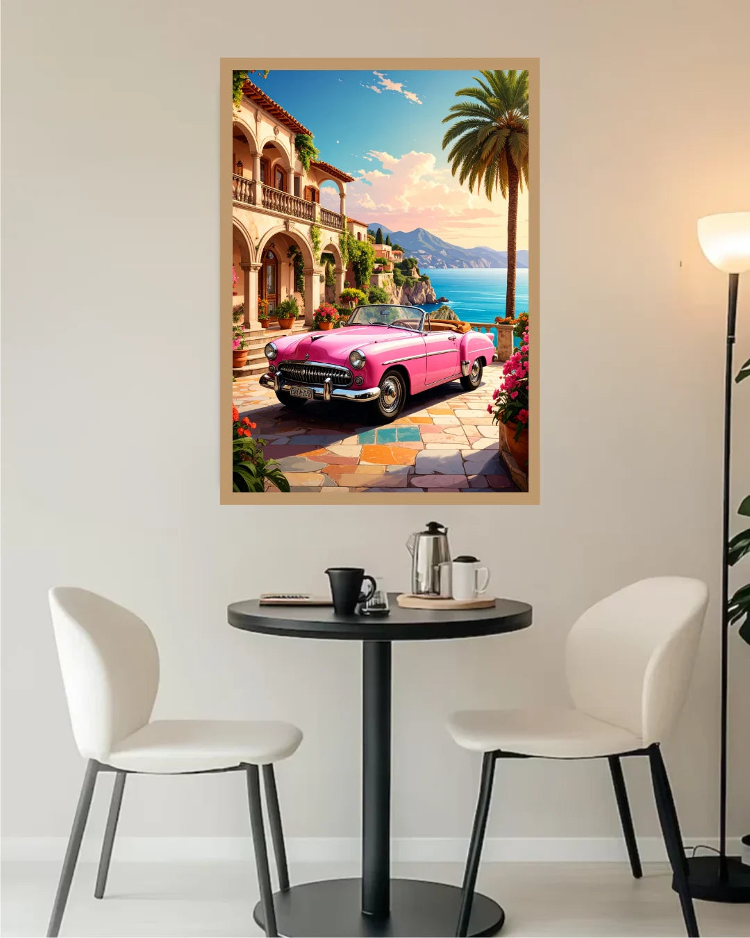 A framed art print depicting a vibrant pink convertible car parked in front of a Mediterranean-style building with a palm tree and blue sea, displayed on a wall above a small dining table with two chairs.