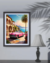 A framed art print depicting a vibrant pink convertible car parked in front of a Mediterranean-style villa by the sea, shown hanging on a light gray wall next to a table lamp with decorative leaf shadows.