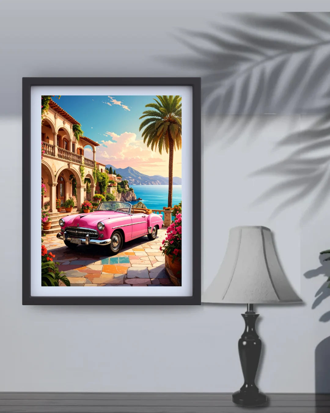 A framed art print depicting a vibrant pink convertible car parked in front of a Mediterranean-style villa by the sea, shown hanging on a light gray wall next to a table lamp with decorative leaf shadows.