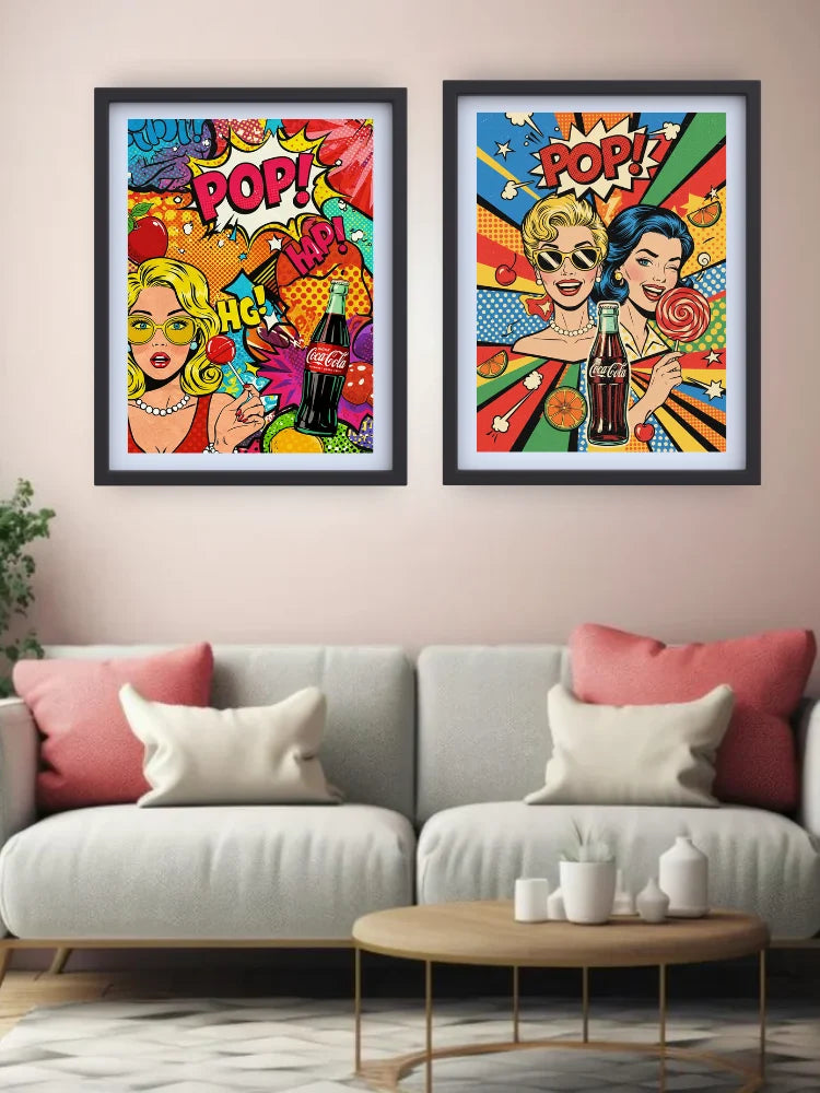 Two framed pop-art posters hanging on a beige wall above a light gray sofa in a living room setting. The posters feature colorful retro comic-style illustrations of women with soda bottles and the word &