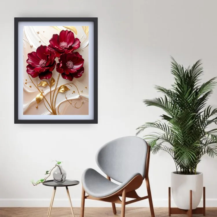 A framed wall art print featuring three deep red flowers with gold stems on a cream background, displayed in a modern room setting above a gray chair and next to a potted plant.