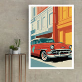 A framed art print featuring a retro 1950s red classic car on a city street, displayed on a textured gray wall next to a small side table with potted plants.