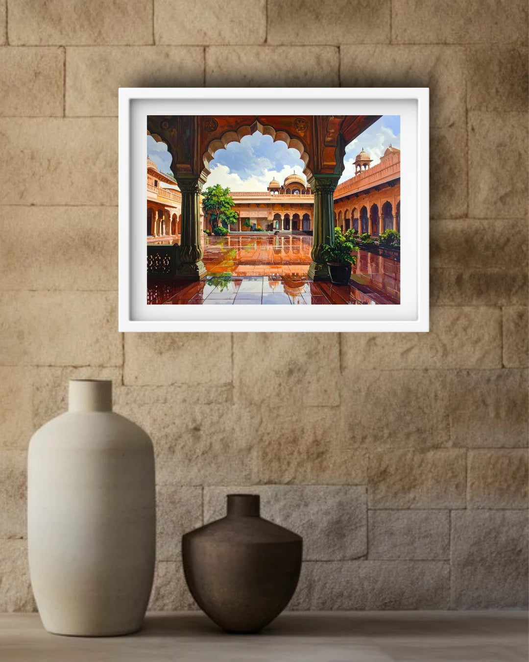 A framed art print depicting a traditional Indian courtyard with arches and a reflecting pool, displayed on a beige stone wall above two ceramic vases.