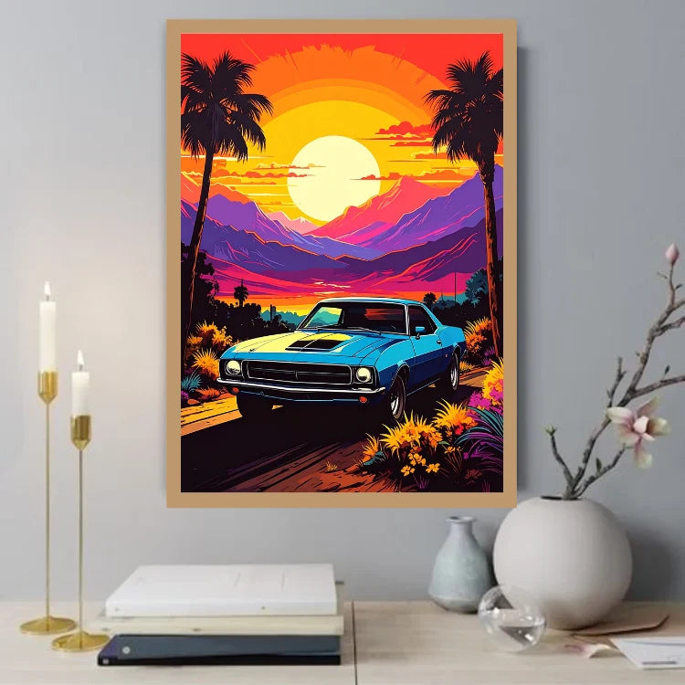 A framed vertical art print featuring a retro blue muscle car on a sunset road with palm trees and mountains, displayed on a tabletop in a styled room setting with candles, books, and vases.