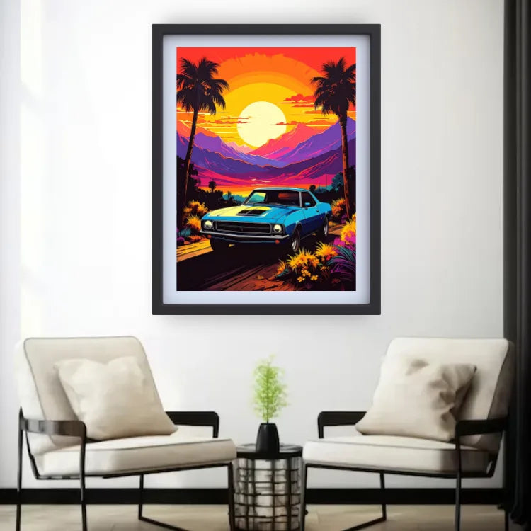 A framed art print featuring a retro blue muscle car on a sunset road trip with palm trees and mountains, displayed on a white wall above two modern beige armchairs.