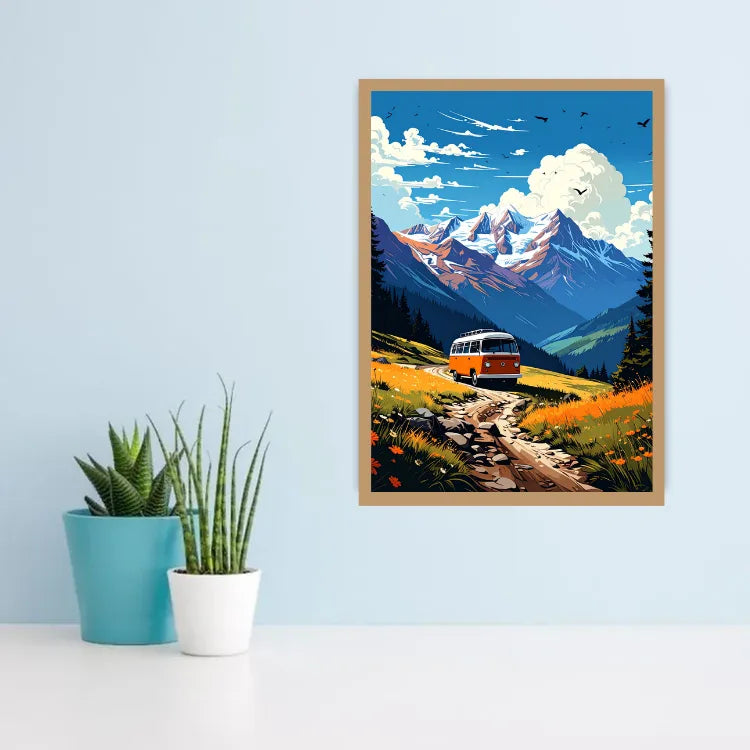 A framed art print depicting a retro orange and white bus traveling down a dirt road through a vibrant mountain valley, hung on a light blue wall above a white surface with two small potted plants.