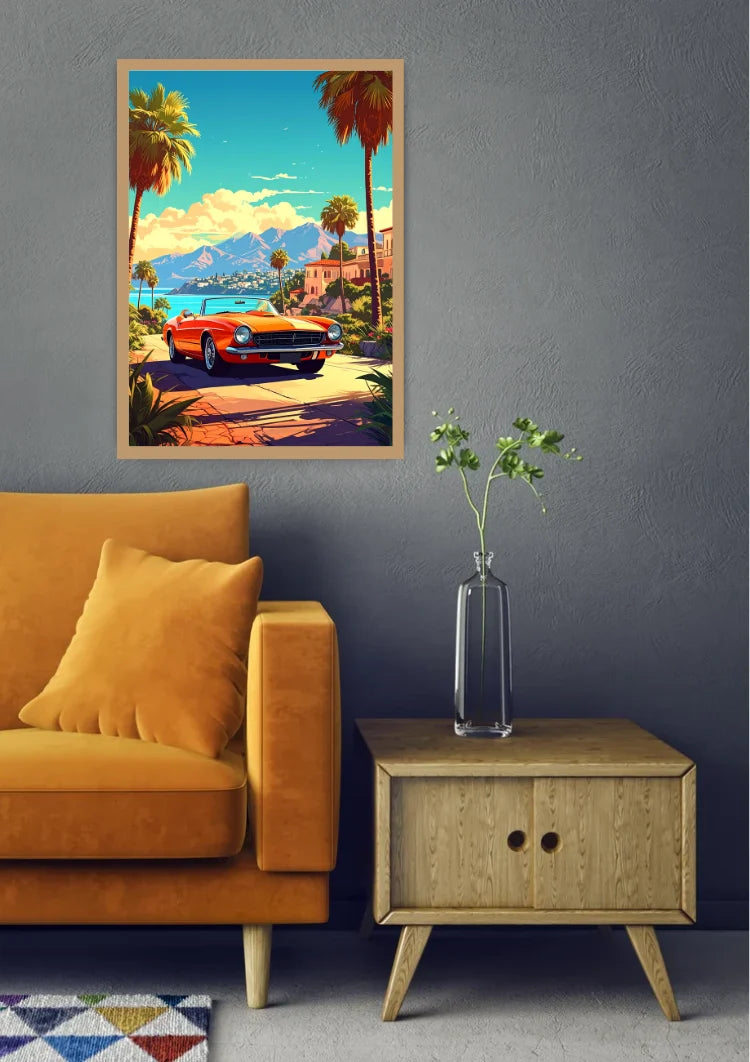 A framed retro-style art print depicting a red classic car on a tropical coastal road with palm trees and mountains, displayed on a dark gray wall above a mustard yellow armchair and a small wooden side table with a glass vase.