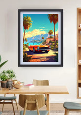 A framed art print featuring a retro orange convertible car on a palm tree-lined road with mountains and a blue sky, displayed on a white wall above a wooden dining table in a modern room setting.