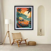 A framed art print featuring a retro orange convertible car on a tropical beach road at sunset, displayed on a cream wall in a styled room with a floor lamp, woven chair, and small vase.