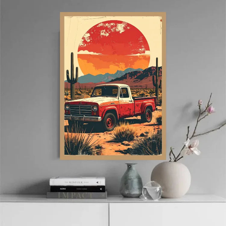 A framed art print featuring a retro red pickup truck in a desert landscape under a large orange-red sun, displayed on a gray wall above a white console table with books and decorative vases.