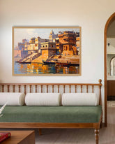 A framed painting of a traditional Indian ghats and river scene hangs on a beige wall above a wooden bench with a green cushion in a styled interior room.