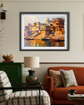 A framed art print depicting a warm-toned painting of a riverfront city with boats and buildings, displayed on a beige wall above a living room setup with an orange sofa, lamp, and chair.