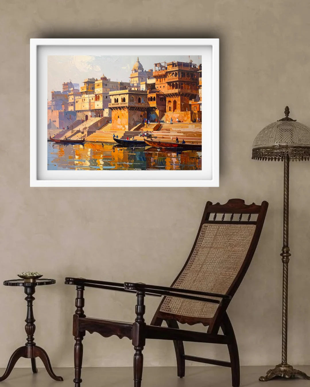 A framed art print depicting a warm-toned painting of a riverfront city with ghats and boats, displayed on a beige wall above a dark wooden lounge chair, with a side table and floor lamp nearby.