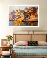A framed wall art print depicting a warm-toned ghats and river scene, displayed above a bed in a styled bedroom setting.