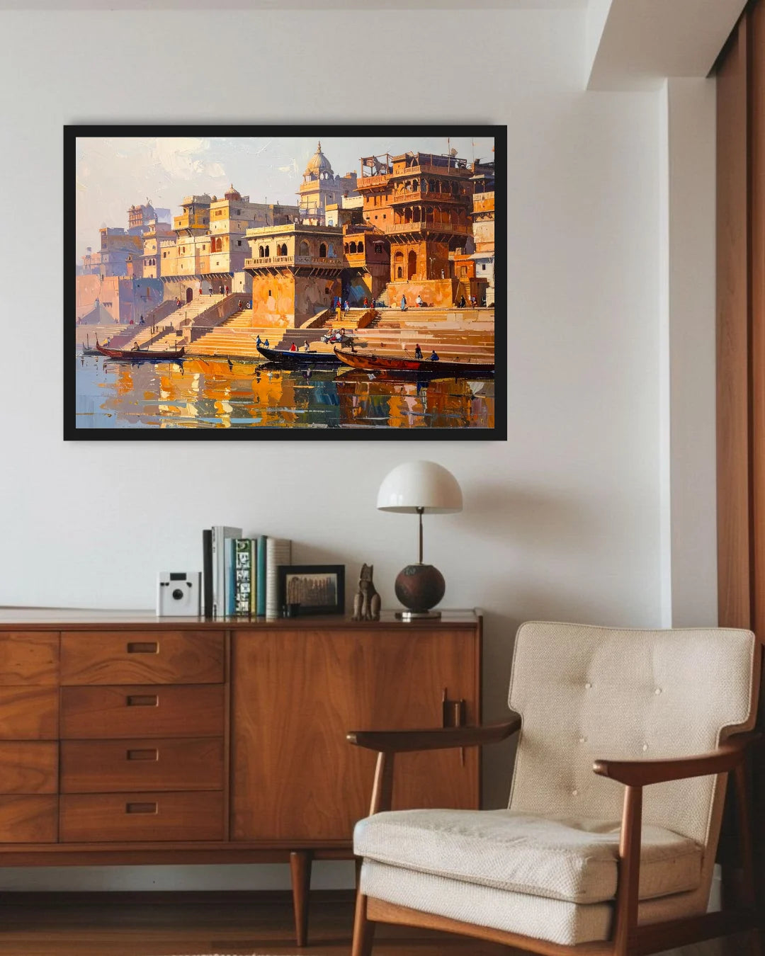A framed painting of a golden-golden river ghat scene with boats and buildings, displayed on a white wall above a wooden sideboard and next to a cream tufted armchair.