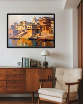A framed painting of a golden-golden river ghat scene with boats and buildings, displayed on a white wall above a wooden sideboard and next to a cream tufted armchair.