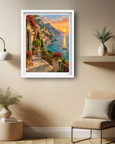 A framed art print depicting a scenic Mediterranean coastal town at sunset, hung on a beige wall in a modern living room setting with a chair, side table, and plants.