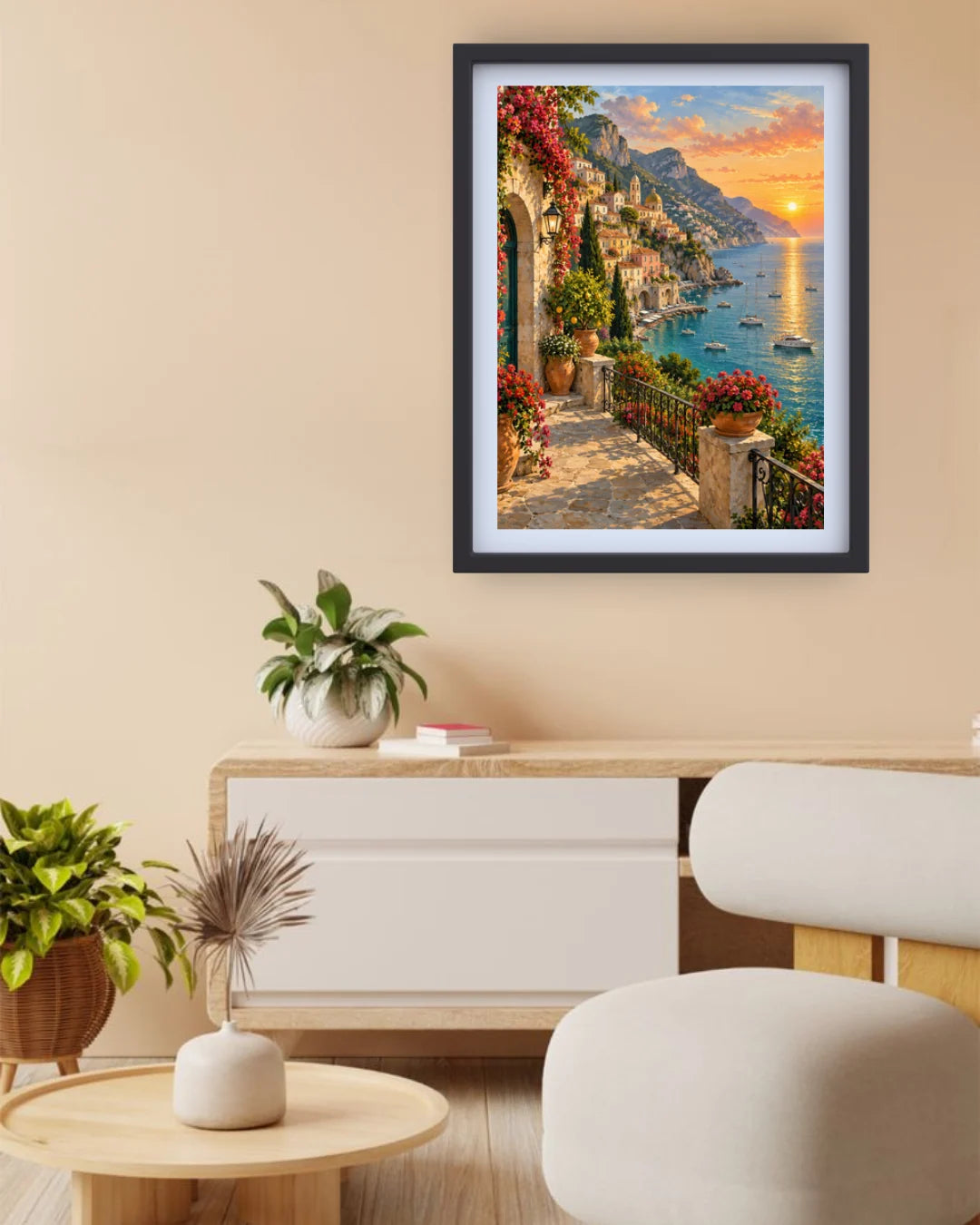 A framed vertical art print depicting a vibrant Mediterranean coastal town at sunset, hung on a beige wall above a modern light wood and white sideboard with potted plants.