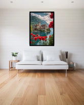 A framed vertical art print depicting a scenic mountain lake with a historic stone church and vivid red flowers in the foreground, displayed on a white paneled wall above a modern white sofa in a bright living room.