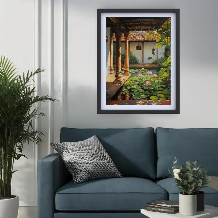 A framed art print depicting a traditional courtyard pond with lily pads and pink flowers, styled on a white wall above a blue sofa in a living room setting.