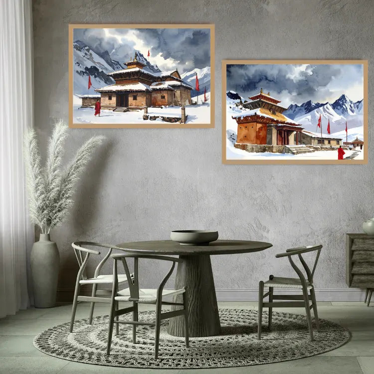 Two framed landscape paintings of snow-covered mountains and traditional buildings displayed on a textured gray wall above a round dining table with chairs in a modern styled room.