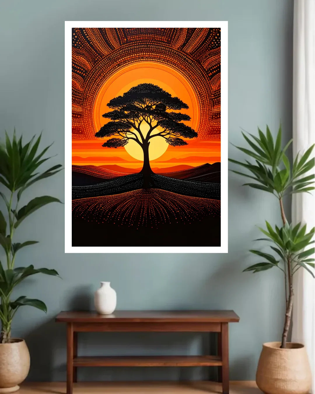 A framed wall art print featuring a stylized tree silhouette against a glowing orange and yellow circular sunset, displayed on a blue-gray wall above a wooden console table with potted plants.