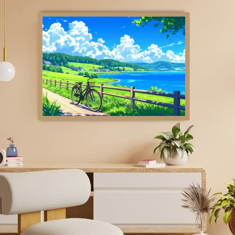 A framed landscape art print depicting a sunny lakeside scene with green fields, blue sky, white clouds, and a bicycle, displayed on a beige wall above a wooden console table in a modern room setting.