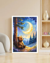 A vertical framed art print featuring a cozy illustration of a teddy bear sitting under a glowing crescent moon in a starry night sky, displayed leaning against a beige wall on a light wood floor next to a chair and shelving.