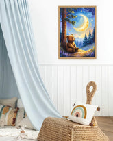 A framed art print depicting a cozy night scene with a teddy bear sitting under a glowing crescent moon in a forest, displayed on a white paneled wall in a styled children&