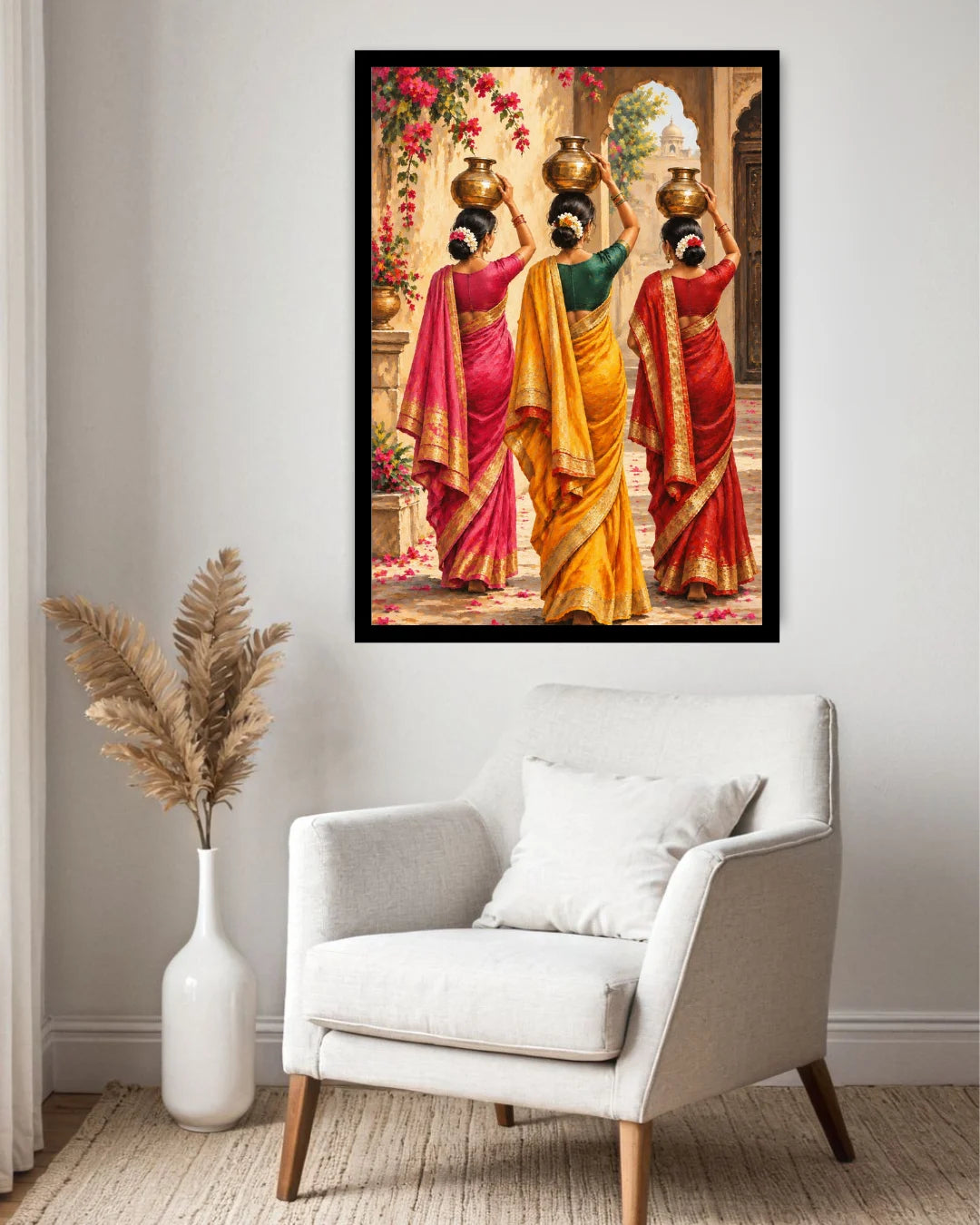 A framed wall art print showing three women in colorful traditional sarees carrying brass pots, displayed above a light gray armchair in a styled room with a white vase of dried grass.