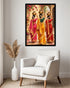 A framed wall art print showing three women in colorful traditional sarees carrying brass pots, displayed above a light gray armchair in a styled room with a white vase of dried grass.