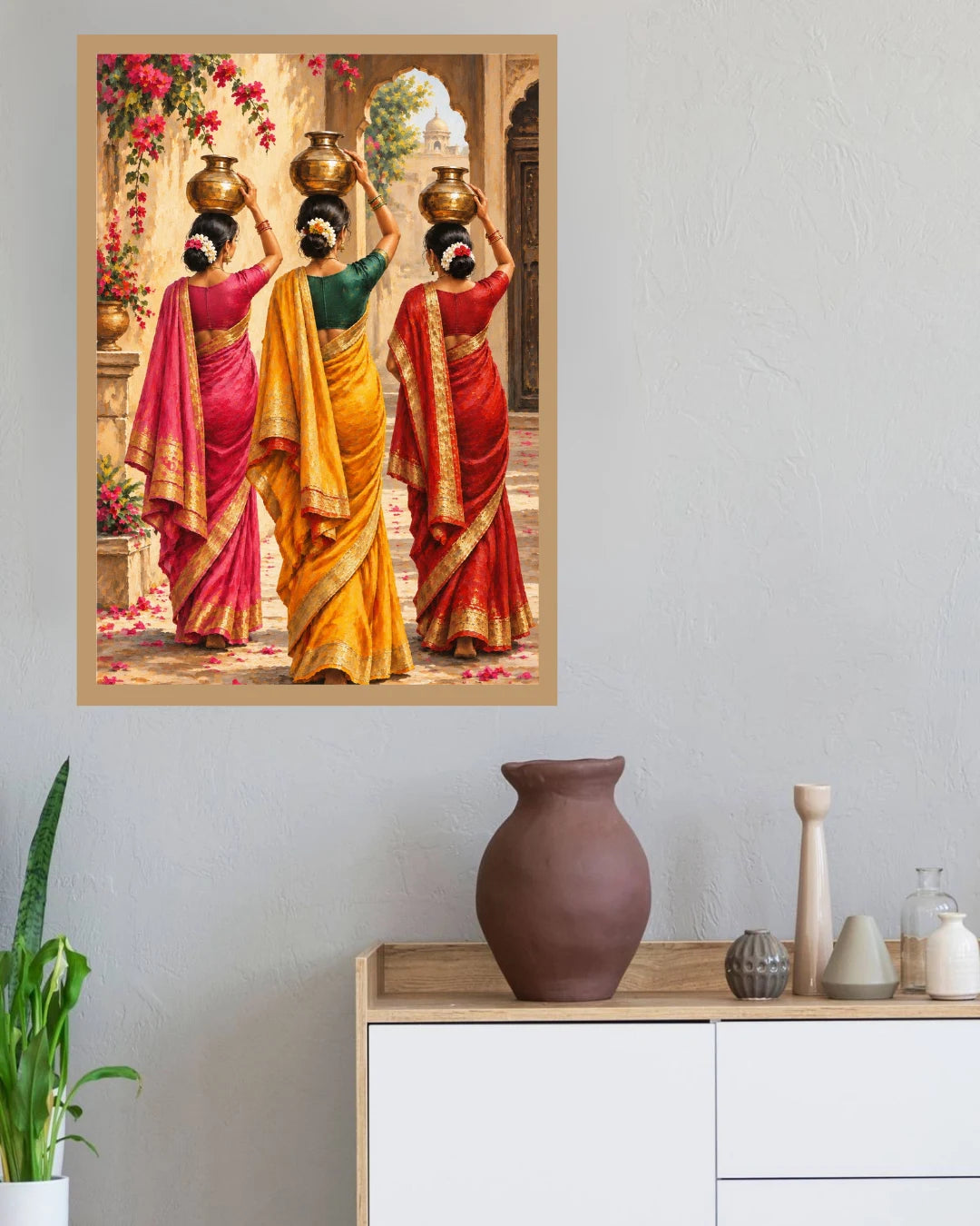 A framed art print depicting three Indian women in colorful sarees carrying brass pots, displayed on a light gray wall above a wooden cabinet with decorative vases and a potted plant.