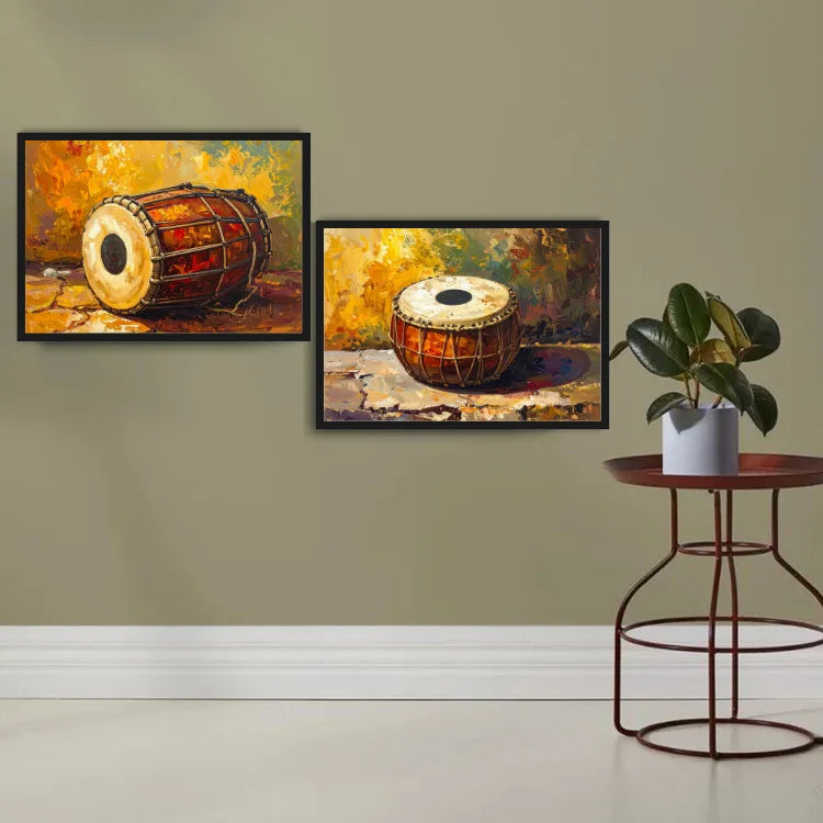 Two framed paintings of traditional Indian drums displayed on a beige wall above a white baseboard, with a small round side table and potted plant on the right.
