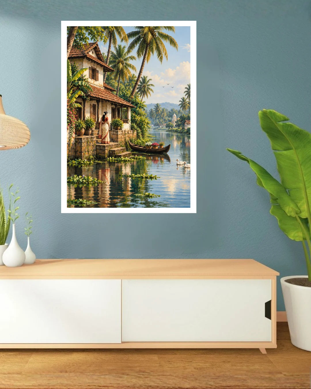 A framed wall art print depicting a tropical lakeside scene with palm trees, a house, boats, and distant hills, displayed on a blue wall above a light wood console table in a styled interior.