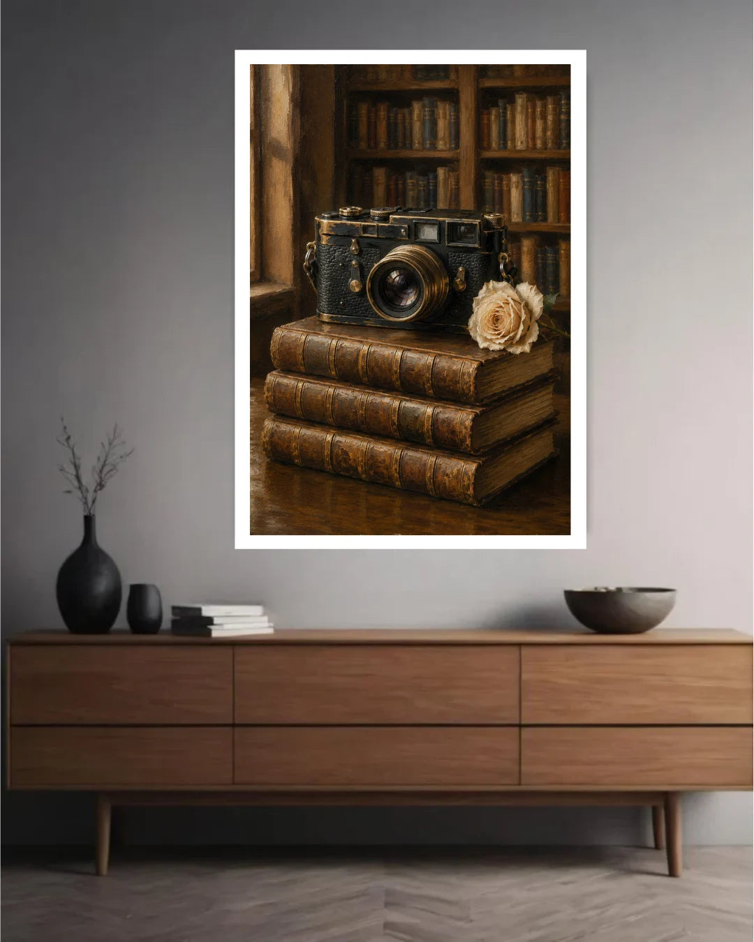 A framed wall art print depicting a vintage camera resting on stacked antique books with a dried flower, displayed above a mid-century modern wooden sideboard in a gray living room setting.