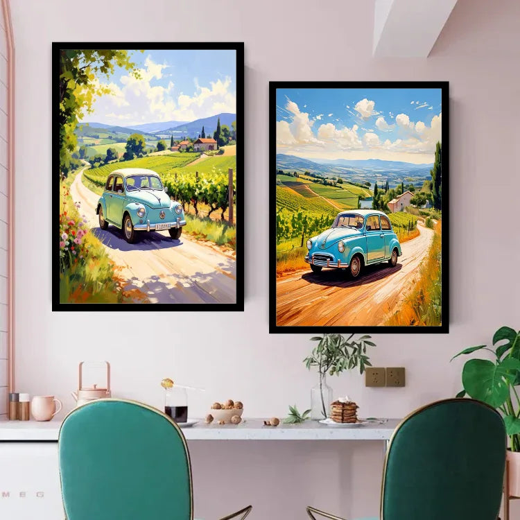 Two framed art prints depicting a light blue vintage car on a winding dirt road through a lush green vineyard landscape, displayed on a light pink wall above a dining table with green chairs.