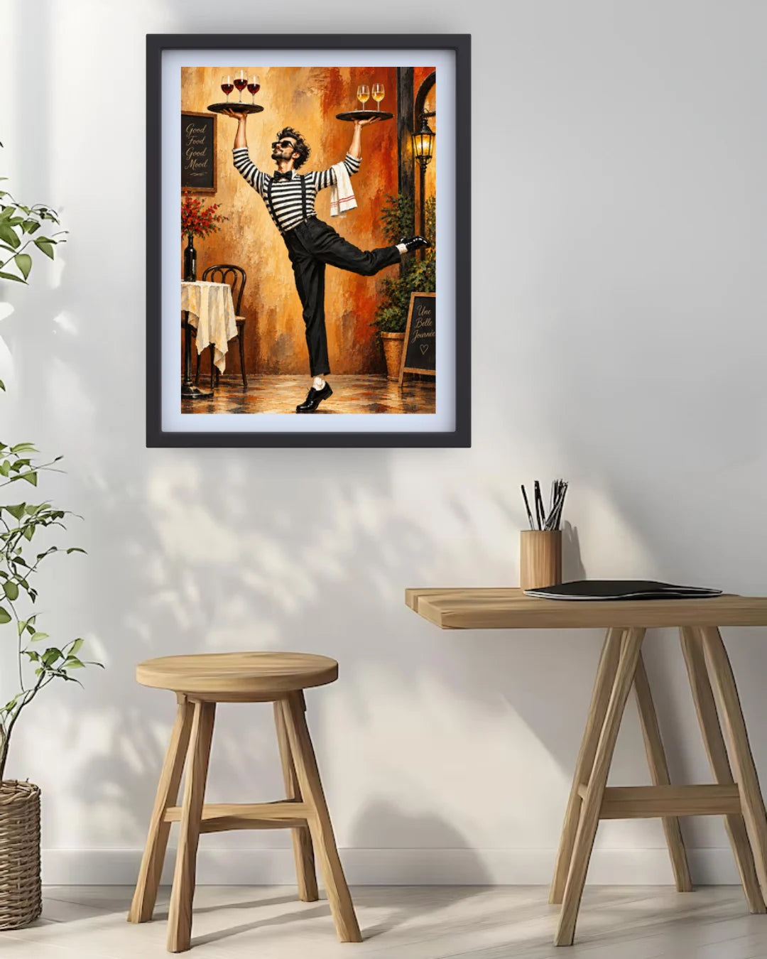 A framed art print featuring a whimsical painting of a woman in a striped shirt balancing wine glasses on trays in a warm-toned restaurant, displayed on a white wall above a wooden table and stool.
