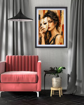 A framed art print depicting a woman holding a white mask, styled in a modern living room setting with a pink armchair, gray curtains, and a small potted plant.