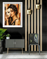 A framed art print depicting a woman holding a white mask, styled in a modern interior setting with a black cabinet, a black chair, gold pendant lights, and a striped wall.