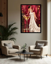 A framed vertical art print depicting a woman in a white gown seated beside a large martini glass, hanging on a beige wall above two cream armchairs and a small black coffee table in a modern living room.