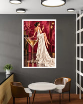 A framed art print featuring a retro-style illustration of a woman in a white gown sitting beside a large martini glass, displayed on a dark gray wall above a small round table and two chairs in a modern interior setting.